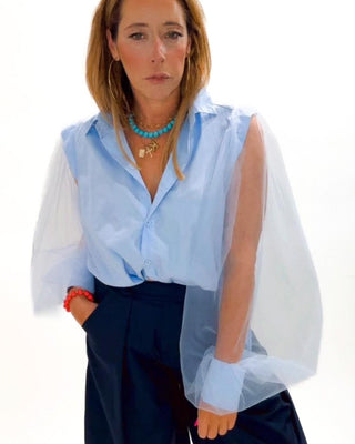 Woman wearing a light blue shirt with sheer sleeves over a white blouse, navy pants, and brown shoes on a white background.